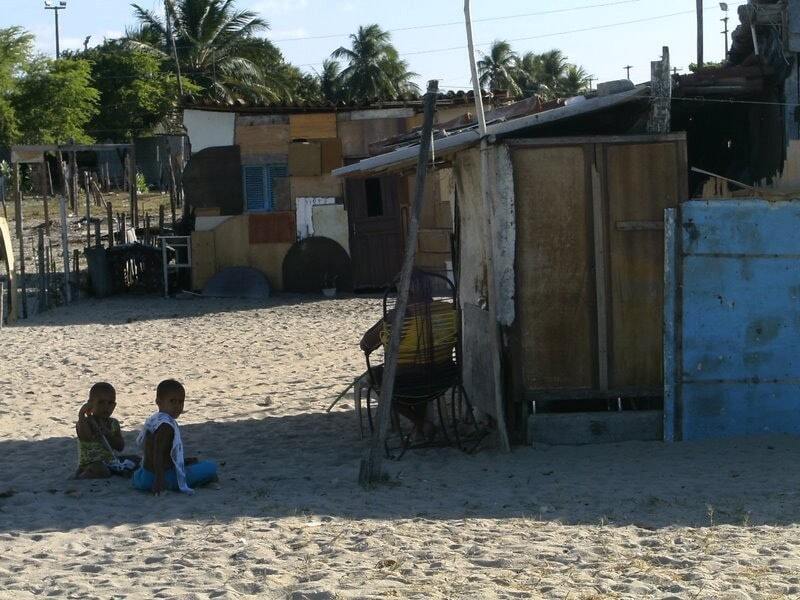 IMAGEM DE UMA COMUNIDADE CARENTE, CRIANÇAS SENTADAS NA AREIA OLHAM CURIOSAS PARA O FOTÓGRAFO