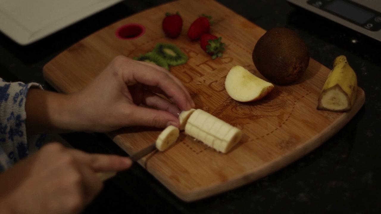 Recheio da Waffle sendo preparado: Kiwi cortado e a banana no professo. Na mesma tábua de cortar morangos e um Kiwi inteiro esperam sua vez